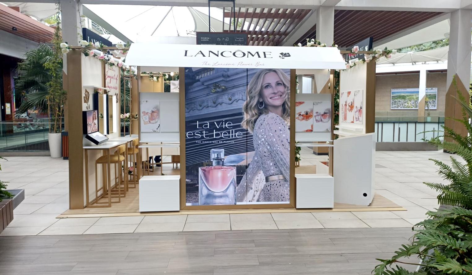 Flower bar Lancôme « La vie est belle » : structure en bois clair, auvent blanc et guirlandes de fleurs.