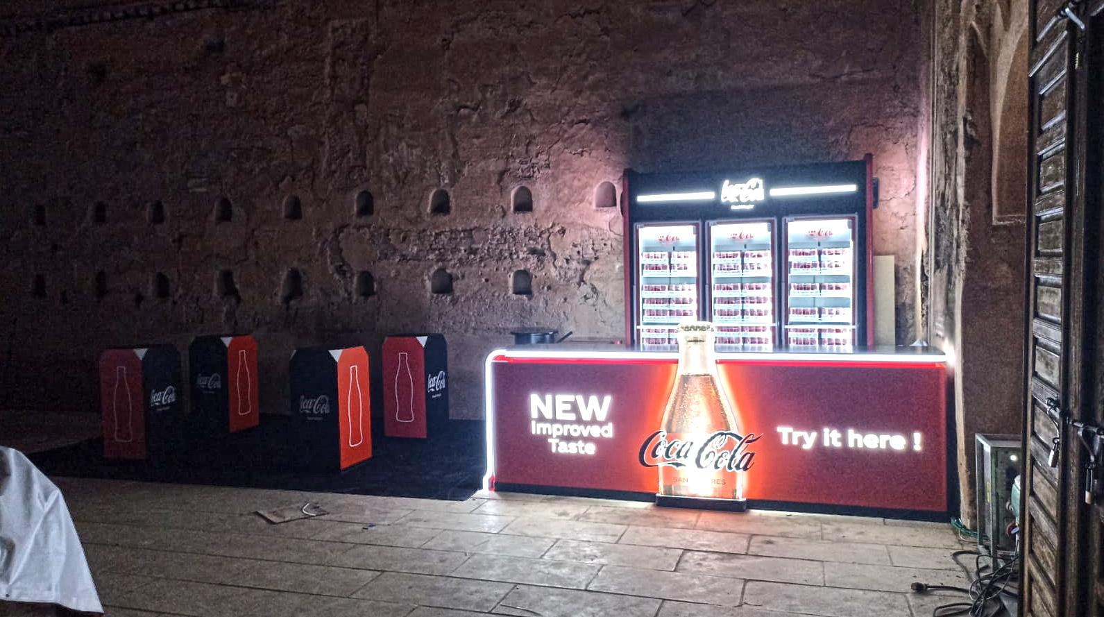 Vue large de nuit : le comptoir lumineux et les assises cubiques Coca-Cola alignées le long du mur.